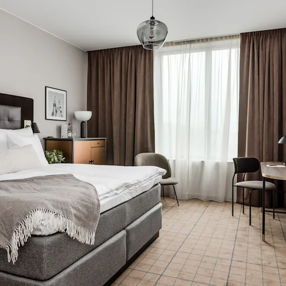 Modern hotelldubbelrum med en stor säng, bruna gardiner, ett litet skrivbord och vägghängd tv.
