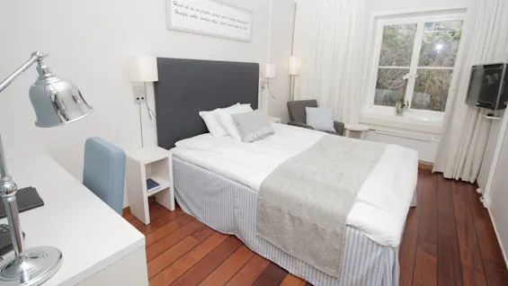 A neatly made bed in a tidy room with furniture, a window, and a text sign on the wall.