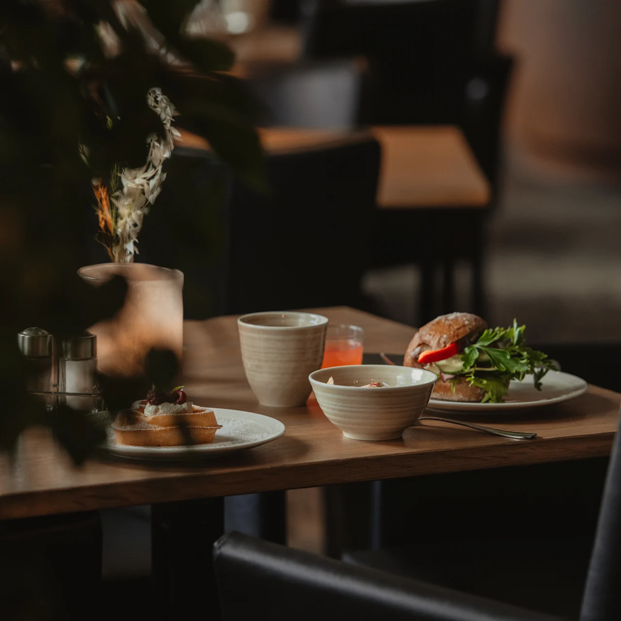A table holds two plates with food; one has a sandwich with greens, the other bread slices. Coffee cups and a juice glass accompany the meal in a cozy restaurant setting.