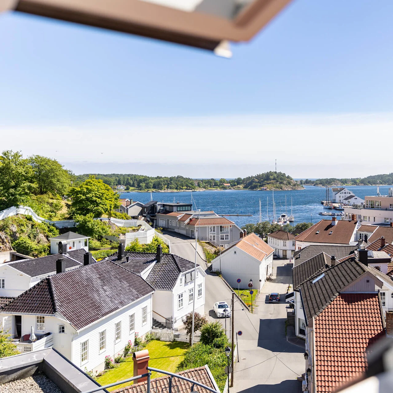 A coastal town with white houses, red roofs, a street, and a car, framed by an open window, overlooking a bay under a clear sky.