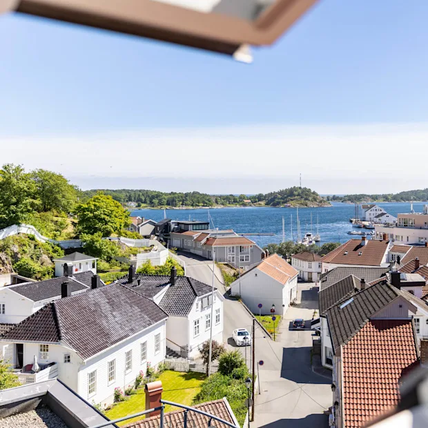 A coastal town with white houses, red roofs, a street, and a car, framed by an open window, overlooking a bay under a clear sky.