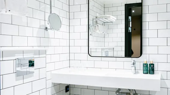 Et hvidt keramisk toilet står i et moderne badeværelse med hvide fliser og et rektangulært spejl over en vægmonteret vask. Håndklæder ligger på en metalhylde, og gulvet er gråt.