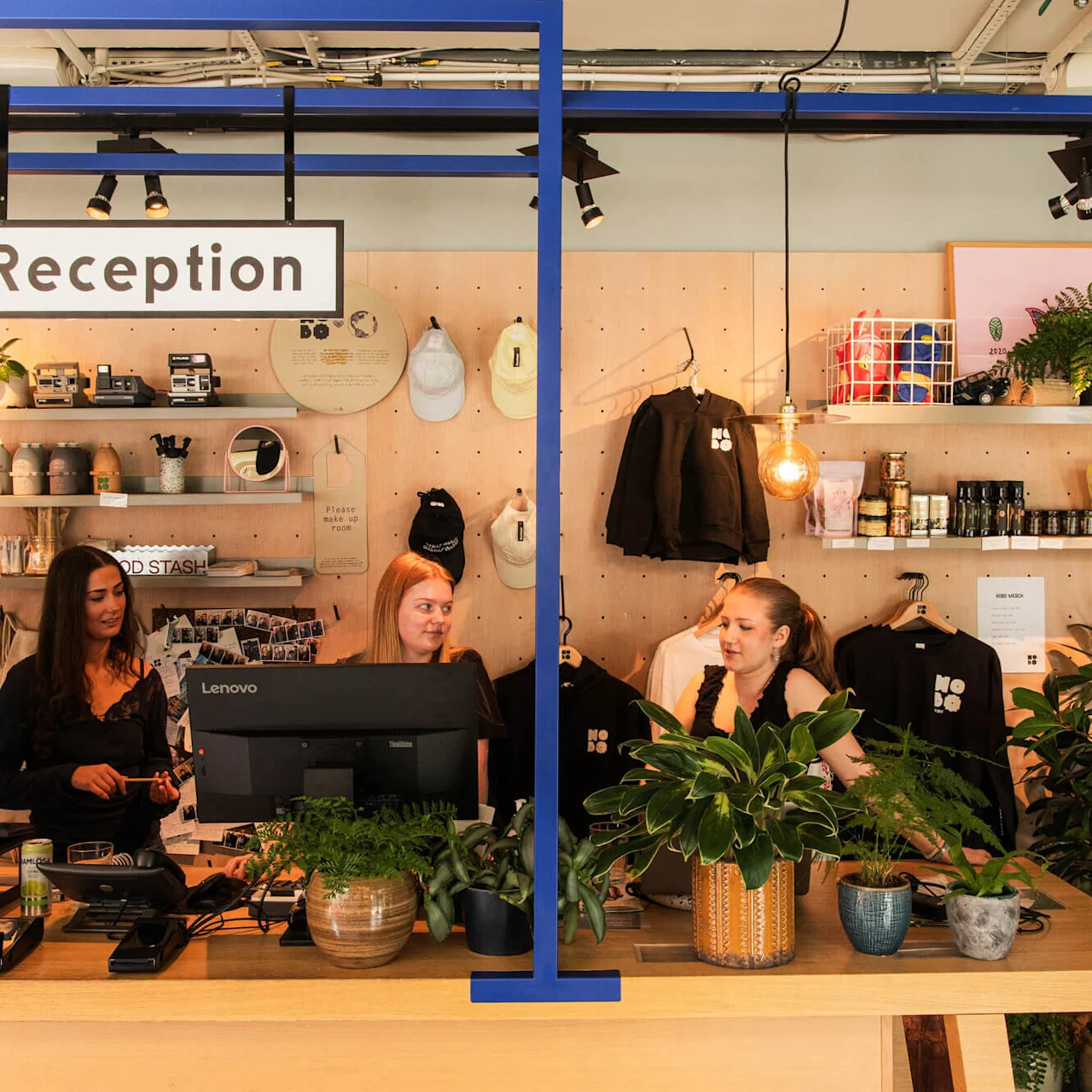 Reception desk with three people working behind computers amidst potted plants. Shelves display merchandise and snacks. A sign reads "Reception." Warm lighting enhances the welcoming ambiance.