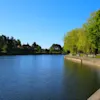 A serene river or lake with a paved path and lush green trees on the right bank, and distant buildings nestled among trees on the left.