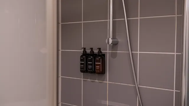 Tre schampoflaskor monterade på en grå kaklad duschvägg med ett silverfärgat duschhuvud i närheten; miljön är ett modernt badrum.