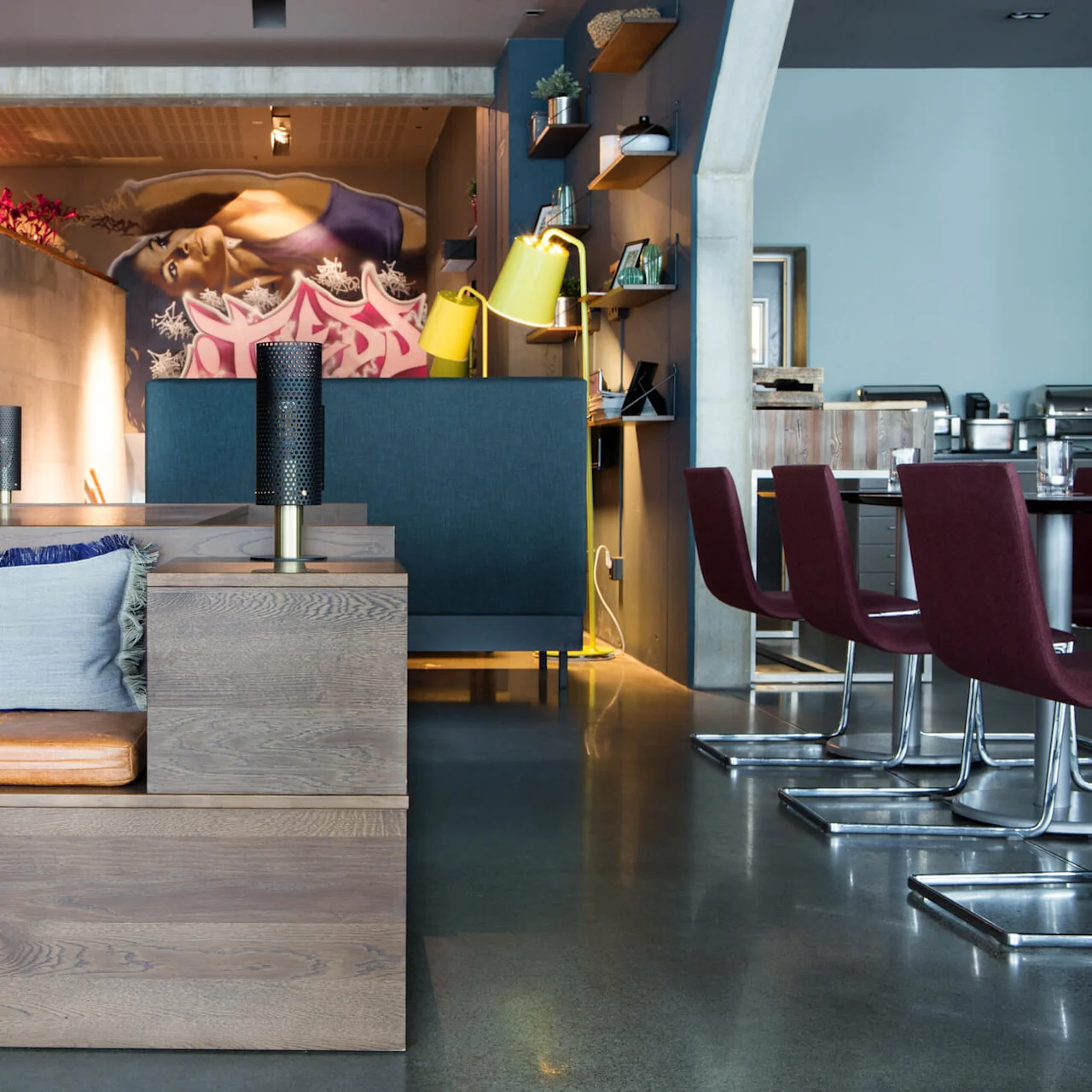 Et moderne restaurantinteriør med trebokser og lærputer i forgrunnen; dekorative lys og hyller pryder veggene, mens trapper fører til et veggmaleri som viser en liggende figur.