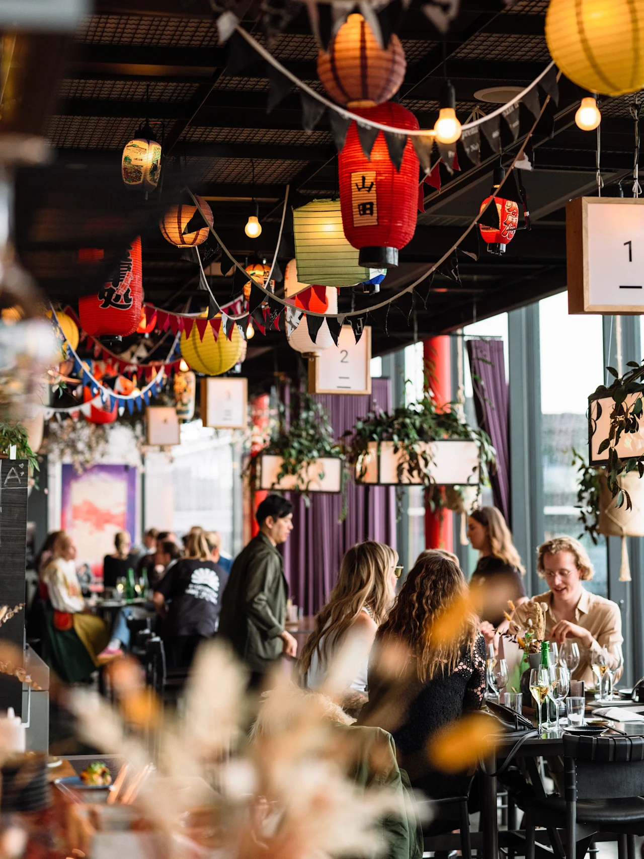 A vibrant restaurant interior with guests dining, decorated with colourful lanterns and plants. Table numbers 1 and 2 are visible.