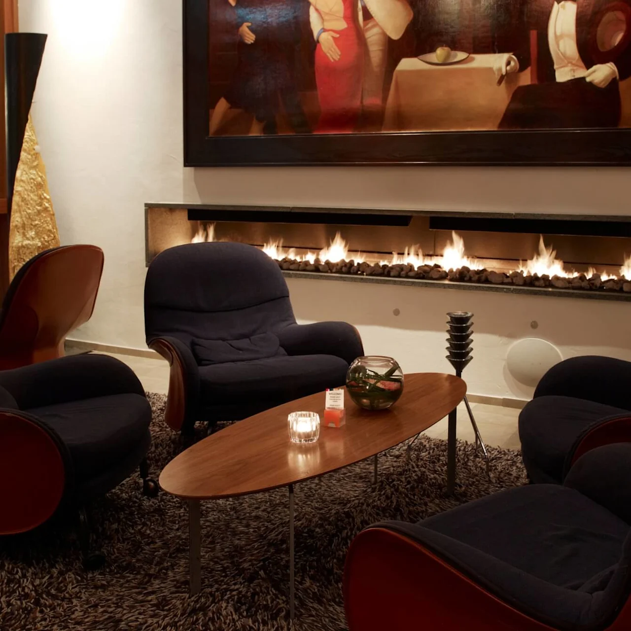 Four dark armchairs encircle a wooden coffee table, adorned with a candle and small vase, in a cozy room with a wall-mounted gas fireplace and an abstract painting above.