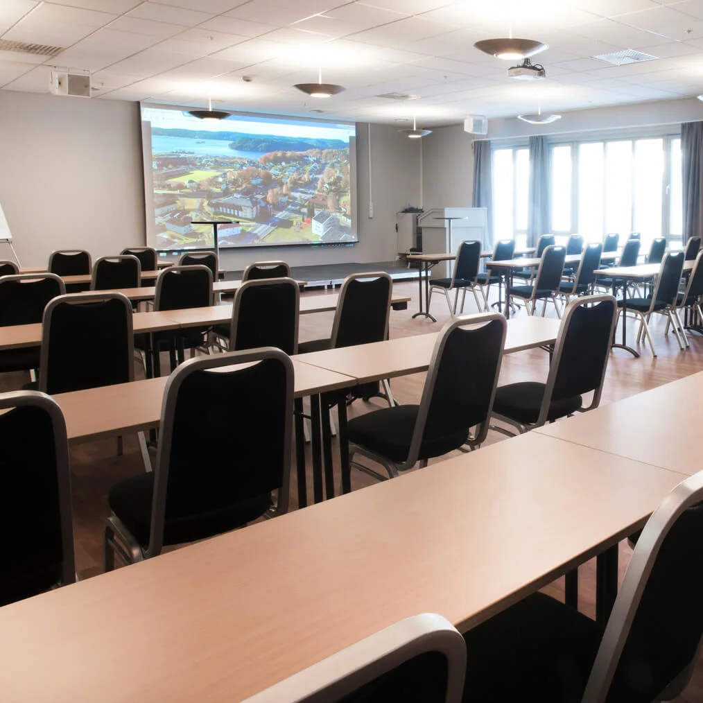 Stolar och bord i rader i ett tomt konferensrum med en projektorskärm på väggen.