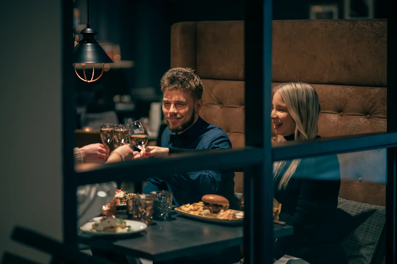Ryhmä ihmisiä nostaa laseja, hymyillen, istuen kodikkaassa ravintolakojussa lautasilla ruokaa, kuten hampurilaisia, ranskalaisia ja juomia pöydällä.
