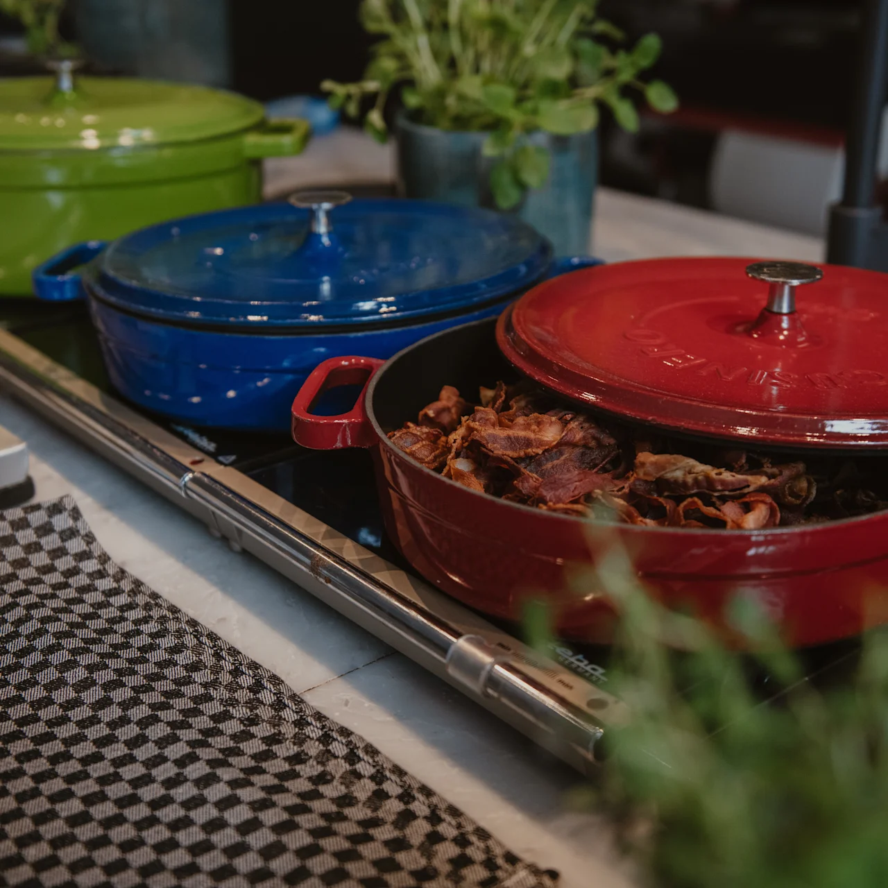 Röd gryta fylld med tillagat kött, delvis öppen på en bänk. Två grytor, en grön och en blå, syns i bakgrunden. Rutig duk och örter omger dem.