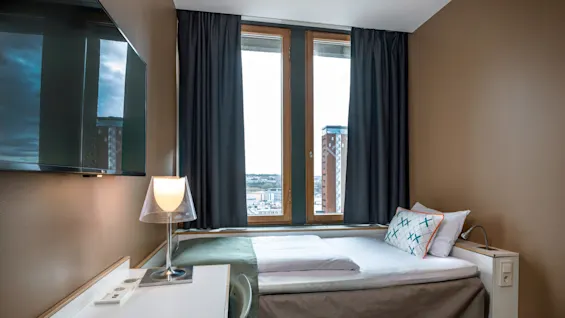 A small, neatly made bed with patterned pillows sits by a large window, next to a desk with a lamp. A TV hangs on a brown wall.