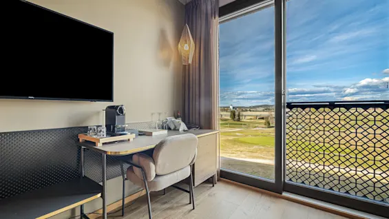 Modern hotelrum skrivbord med en kaffemaskin och stol, bredvid ett stort fönster med utsikt över ett fält och blå himmel. TV monterad på väggen. Ingen text.