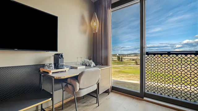 Moderne hotellrom pult med kaffetrakter og stol, ved siden av et stort vindu med utsikt over et felt og blå himmel. TV montert på veggen. Ingen tekst.