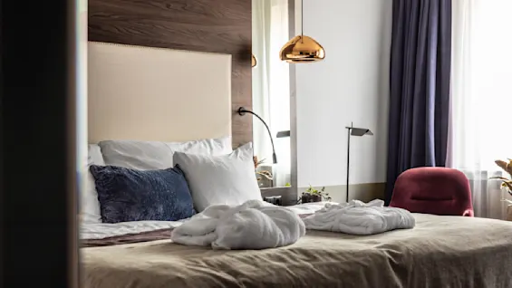 Ett mysigt hotellrum med en prydligt bäddad säng, dekorerad med kuddar och ett par morgonrockar. Mjukt ljus sipprar genom gardiner bredvid en stilfull röd stol. En varm och inbjudande atmosfär fyller rummet.