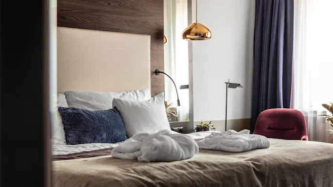 Et koselig hotellrom med en pent oppredd seng, pyntet med puter og noen badekåper. Mykt lys slipper inn gjennom gardinene ved siden av en stilig rød stol. En varm, innbydende stemning fyller rommet.