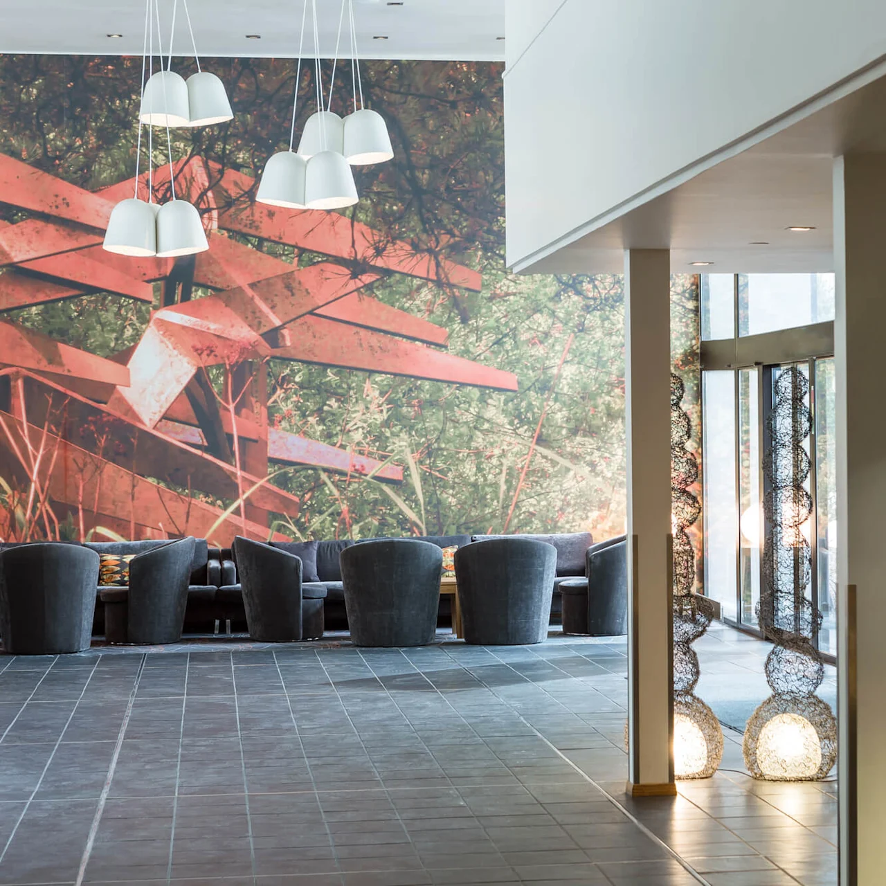 Seating area with dark chairs faces mural of red abstract sculpture and greenery. Decorative hanging lights illuminate above; snack stand with "Lounge 24" sign nearby.