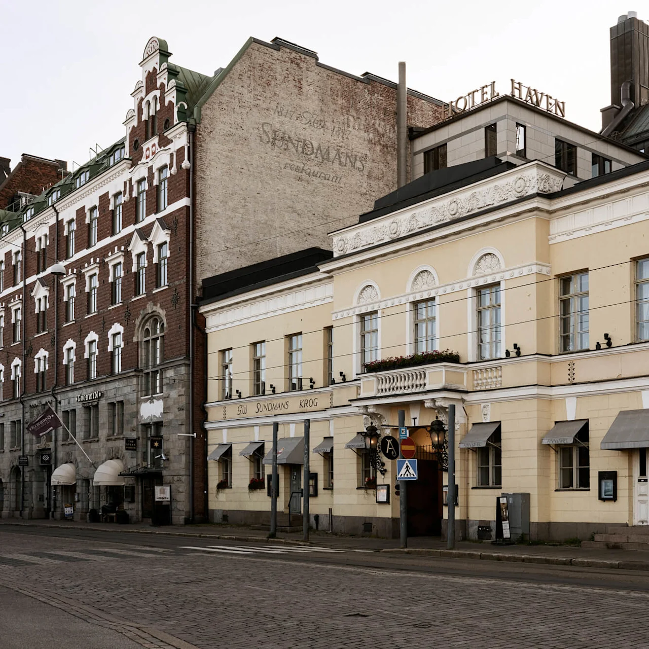 A yellow building features "HOTEL HAVEN" and "RESTAURANT" signage. Neighboring structures are ornate, with arched windows and decorative designs. Street view includes cobblestone road and pedestrian crossing signs.
