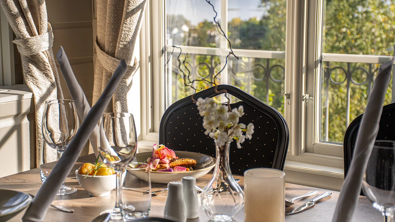 Ett matbord dukat för en måltid, med mat, dryck och blomsterdekor, badat i naturligt ljus från ett fönster.