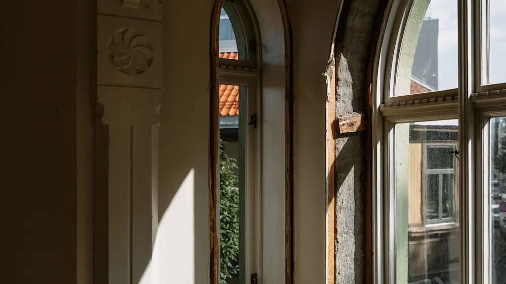 Sollys strømmer ind gennem to buede vinduer i et rum under renovering og kaster dramatiske skygger på væggene.