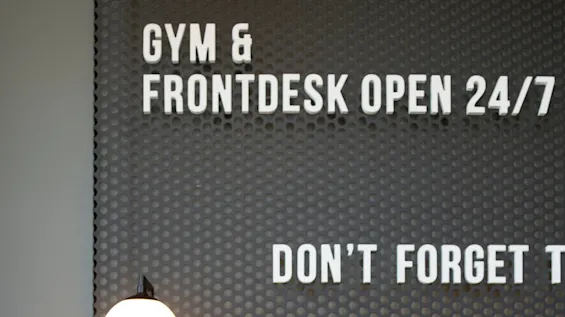 Bed with striped white sheets against a perforated metal headboard. Wall text reads, "GYM & FRONTDESK OPEN 24/7 DON’T FORGET TO." Lamp and outlets are visible.