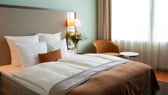 A neatly made bed with white pillows and a brown blanket rests by a window. A lamp, flowers, a chair, and a small table enhance the cozy room.