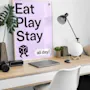 Juliste puupöydällä, jossa teksti "Eat Play Stay all day!" Sisältää noppakuvituksen, ympärillä toimistotarvikkeita kuten kasvi, lamppu ja kannettava tietokone minimalistisessa huoneessa.