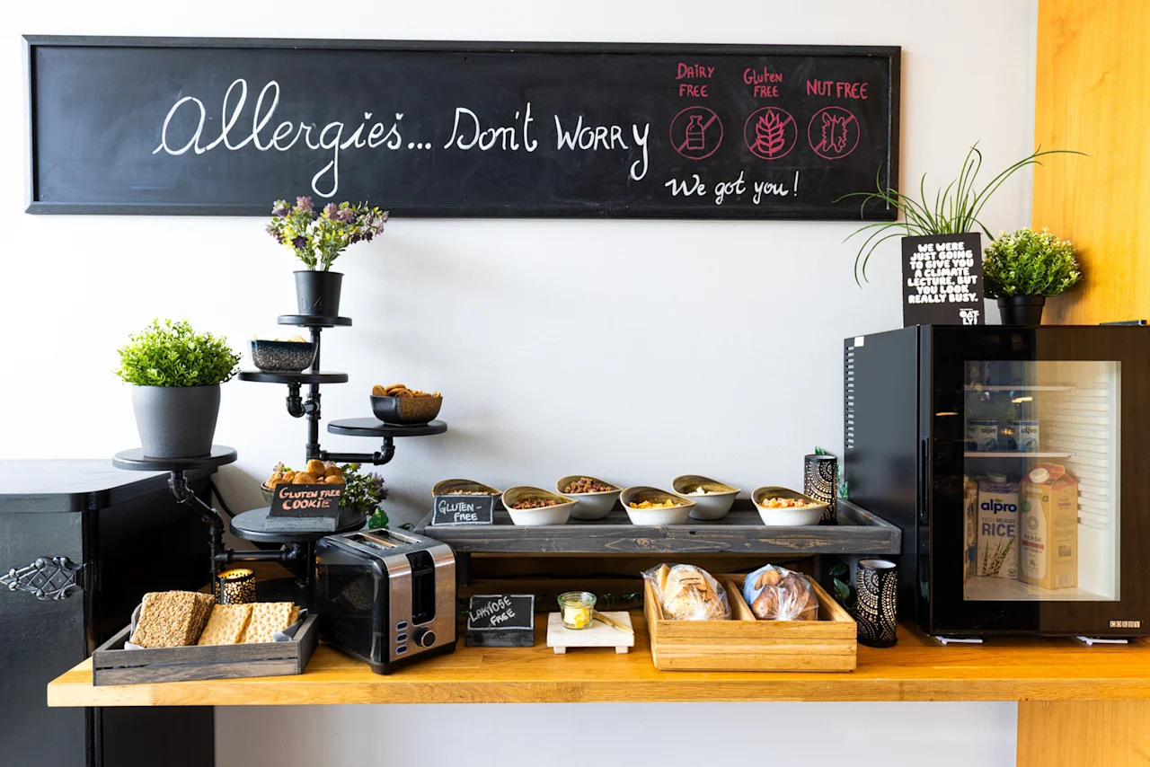 En frokostbuffet har korn, en brødrister, brød og bakverk på en trebenk. På en tavle over står det: "Allergies...Don't Worry. We got you! Dairy free, Gluten free, Nut free."