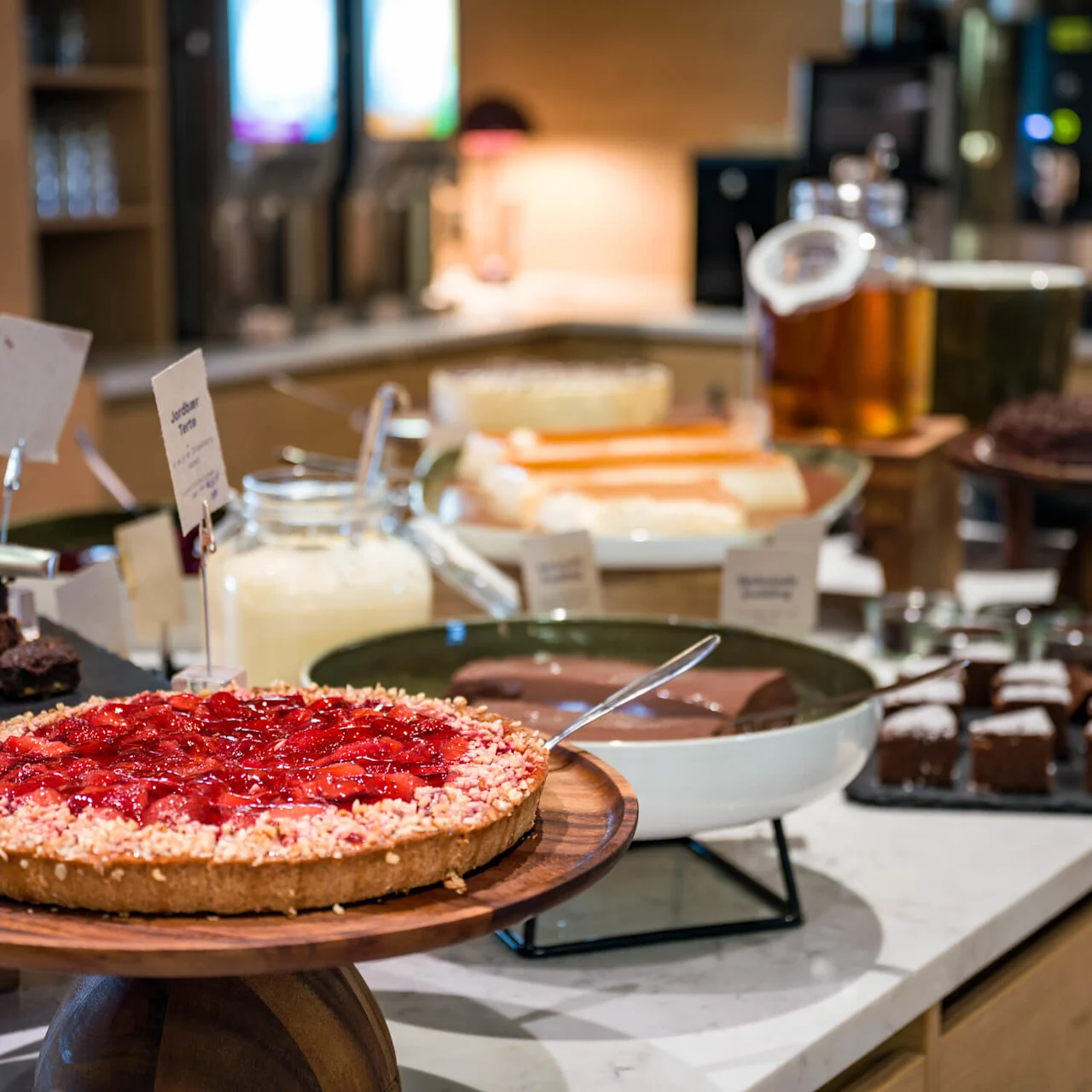 Jordbærterte på en trebordplate, omgitt av forskjellige desserter som brownies og pudding. Plassert i et buffetmiljø med myk belysning.