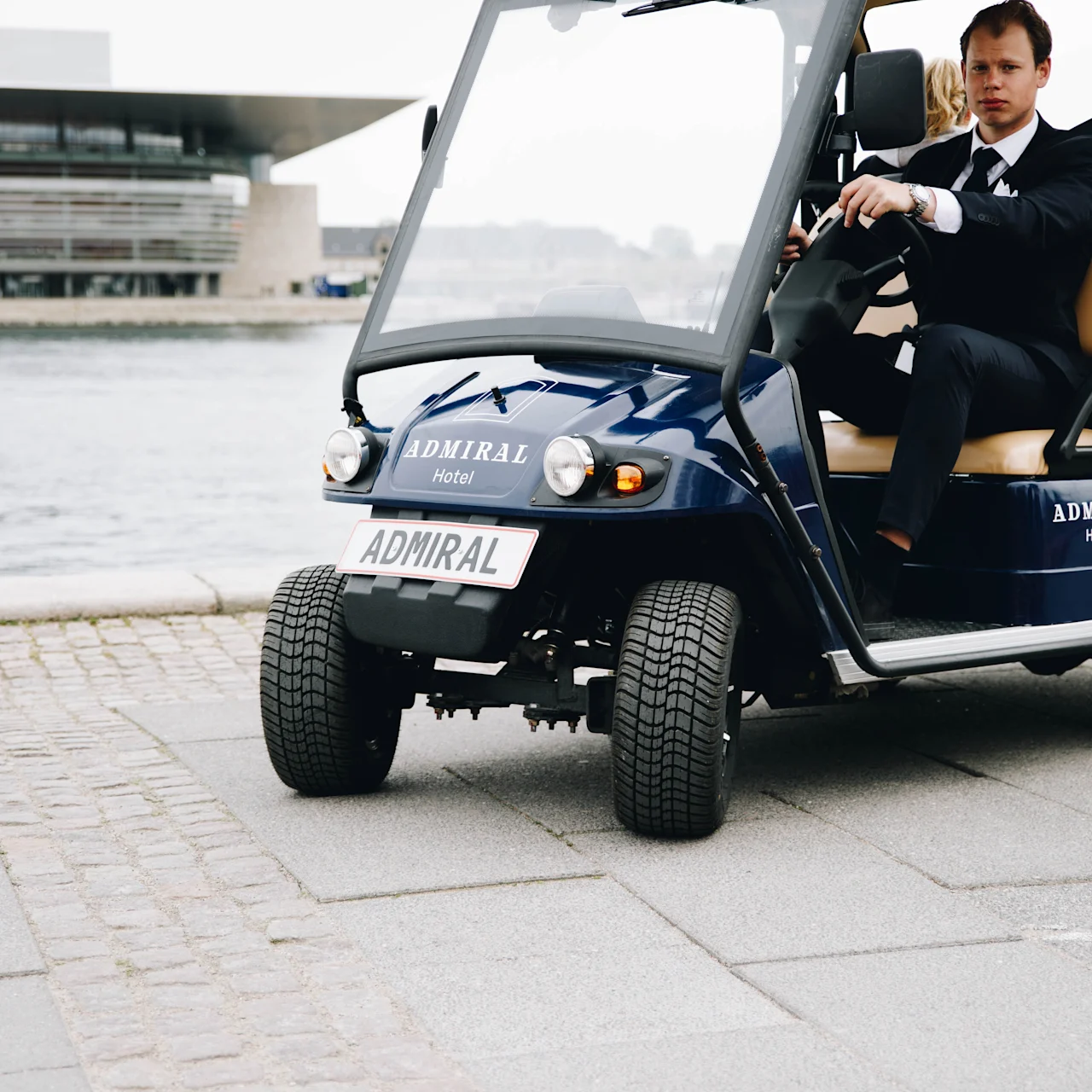 Golf cart driven by a man in a suit beside a waterfront, near a modern building with text "ADMIRAL Hotel" and "ADMIRAL" on the license plate.