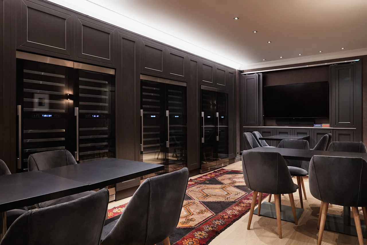 Dark dining tables and chairs are arranged in a restaurant setting. Wine coolers line the wall behind, with a mounted TV in the corner. A decorative carpet accents the floor.