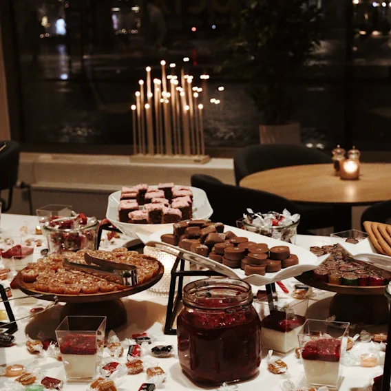 Festive dessert buffet table filled with chocolates, small cakes, mousse cups, and candies, lit by a warm advent candelabra in the background window.
