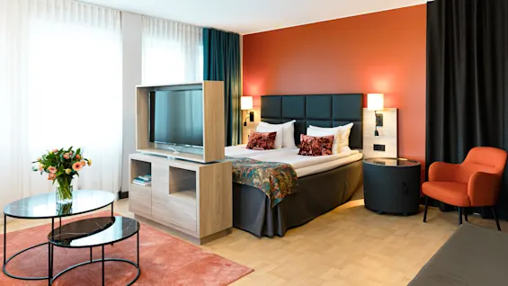 Ett modernt hotellrum har en prydligt bäddad säng med kuddar, sänglampor, en soffstol, en TV på en träställning och runda soffbord, mot eleganta gardiner.