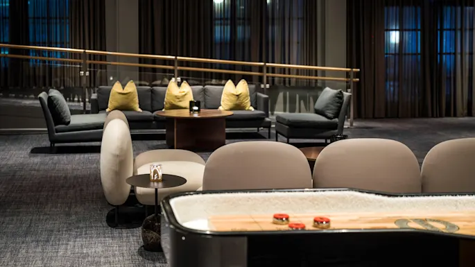 Shuffleboard table, with red pucks, stands in a dimly lit lounge area. Surrounding are modern chairs and a sofa with yellow pillows. Dark curtains and soft lighting create a cozy ambiance.