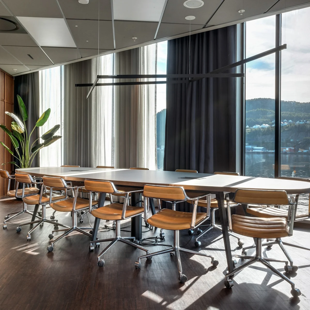 Et langt konferencebord omgivet af læderstole står i et moderne rum med store vinduer. En plante dekorerer hjørnet, og bjerge er synlige udenfor.