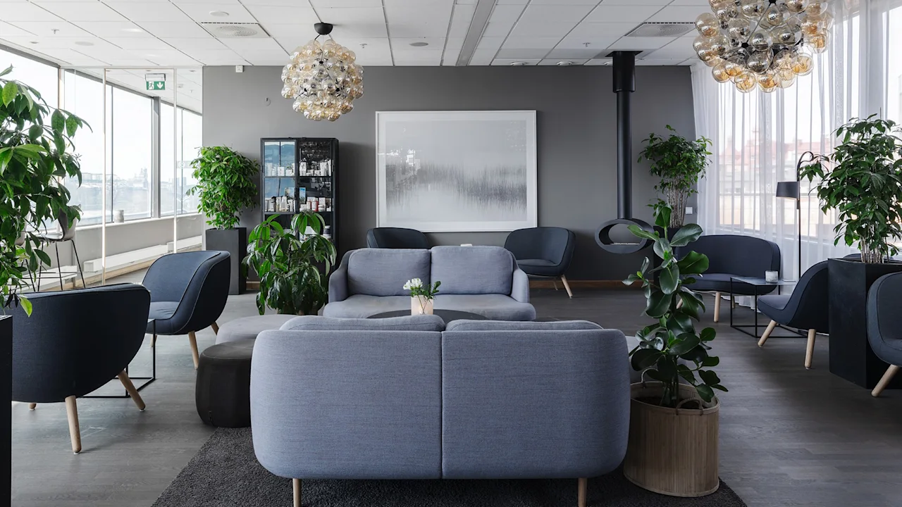 Komfortable grå sofaer står vendt mot hverandre i en moderne lounge. Frodig grønne planter omgir, med store vinduer som gir naturlig lys. Stilfulle taklamper og minimalistisk interiør fullfører rommet.