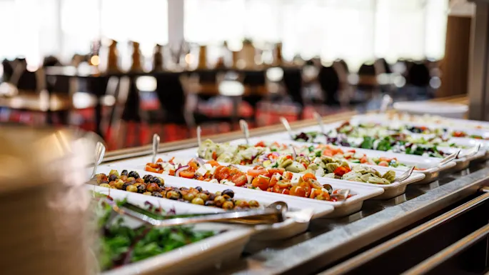 Et buffetbord viser forskjellige fargerike salater i hvite brett med serveringsskjeer, i en restaurantinnstilling med bord og uklar stoler i bakgrunnen.