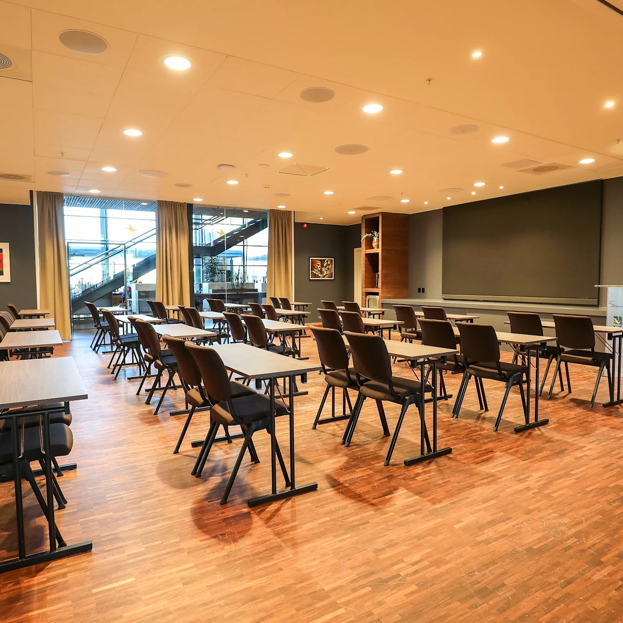 Rader av svarte stoler vendt mot bord på et tregulv i et godt opplyst konferanserom; malerier pryder veggene, og store vinduer avslører en trapp utenfor.