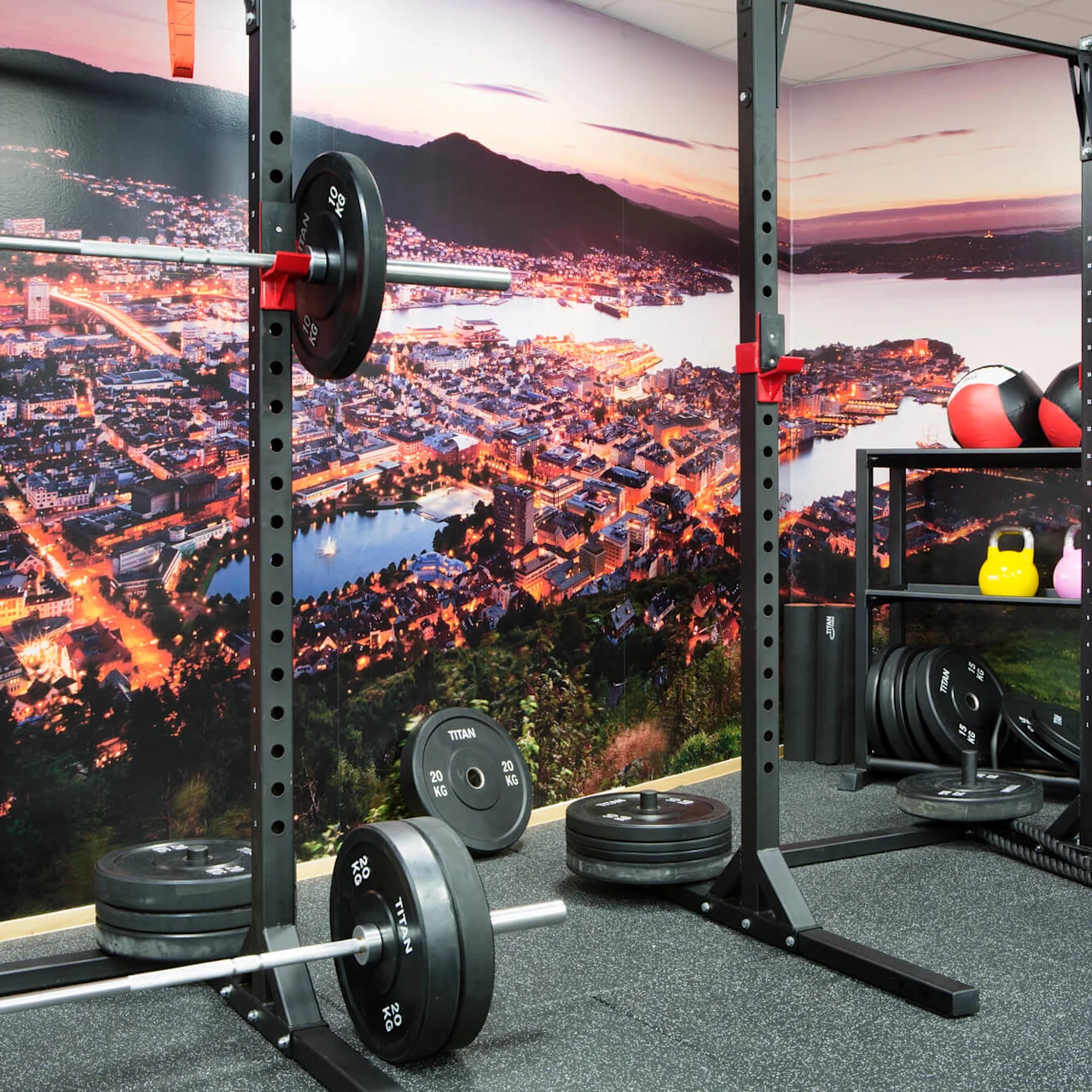 A weightlifting gym features a barbell with weights and various kettlebells. The wall displays a panoramic cityscape at sunset, creating a vibrant atmosphere.