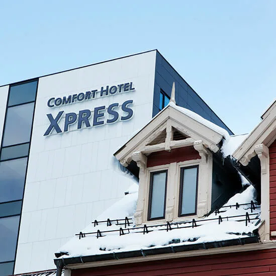 A modern building labeled "COMFORT HOTEL XPRESS" stands beside a snow-covered, red wooden house with a pointed roof in an urban setting.