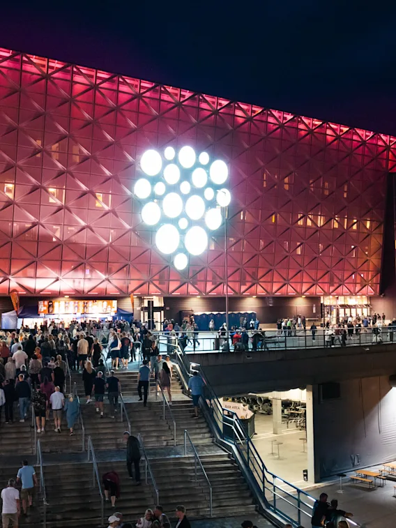 A large crowd of people walking up stairs towards a brightly lit arena at night. Arena.