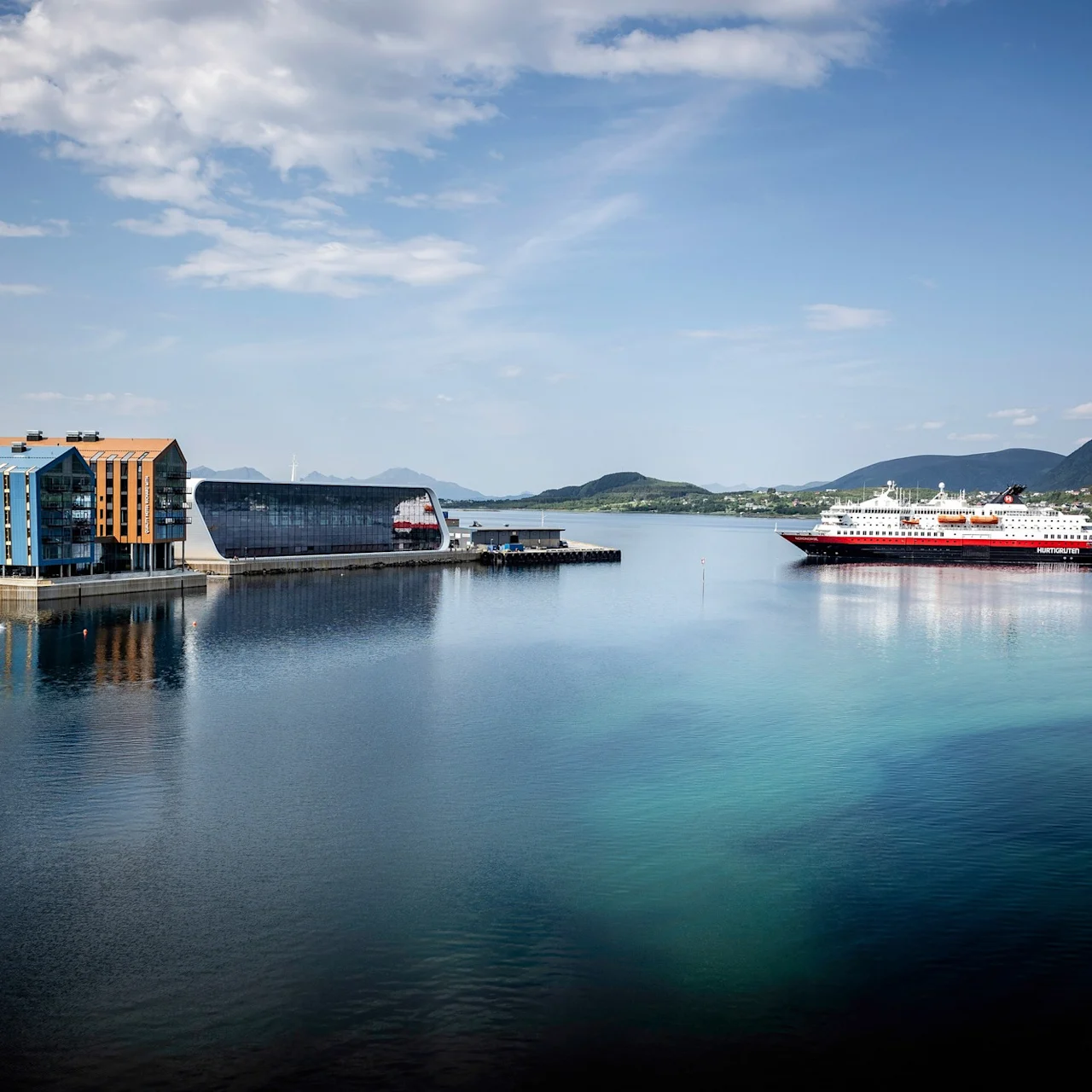 En stor færge mærket "Hurtigruten" sejler over en rolig blå fjord. Farverige bygninger og bjerge omgiver kysten under en klar himmel.