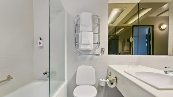 Toilet under håndklædestativ med hvide håndklæder, ved siden af et badekar med glasvæg. Håndvask med to glas og stort spejl til højre.