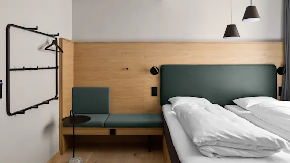 En prydligt bäddad säng med vita lakan står bredvid en stoppad bänk mot en träpanel. En minimalistisk klädhängare och två hängande lampor kompletterar det moderna, luftiga rummet.