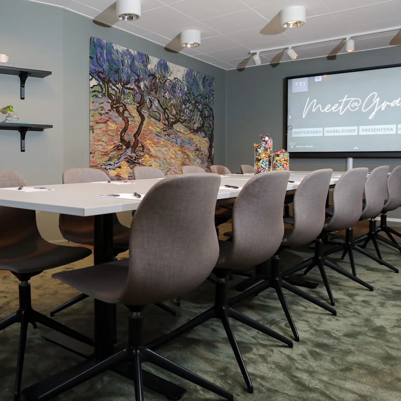 Meeting room table with gray chairs, a large screen displaying "Meet & Greet," and a vibrant painting on the wall. Shelves with plants and bottles decorate the room.