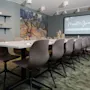 Meeting room table with gray chairs, a large screen displaying "Meet & Greet," and a vibrant painting on the wall. Shelves with plants and bottles decorate the room.