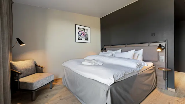 En seng med hvitt sengetøy og puter er plassert i et moderne rom. Ved siden, en komfortabel stol og en gulvlampe forsterker den innbydende atmosfæren.