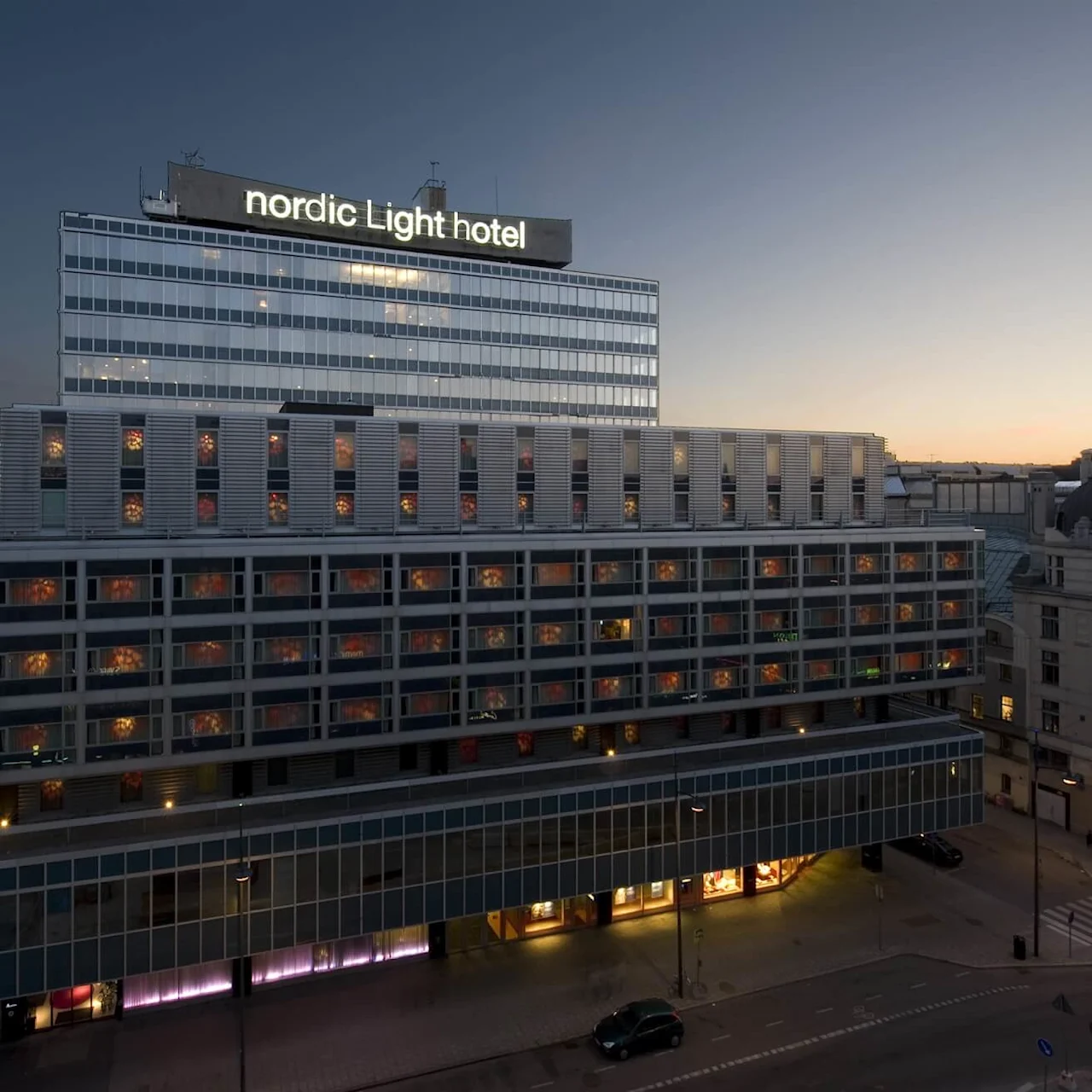 A building with "nordic Light hotel" sign displays lit windows; it's dusk, creating a serene urban setting with a few parked cars on the street below.