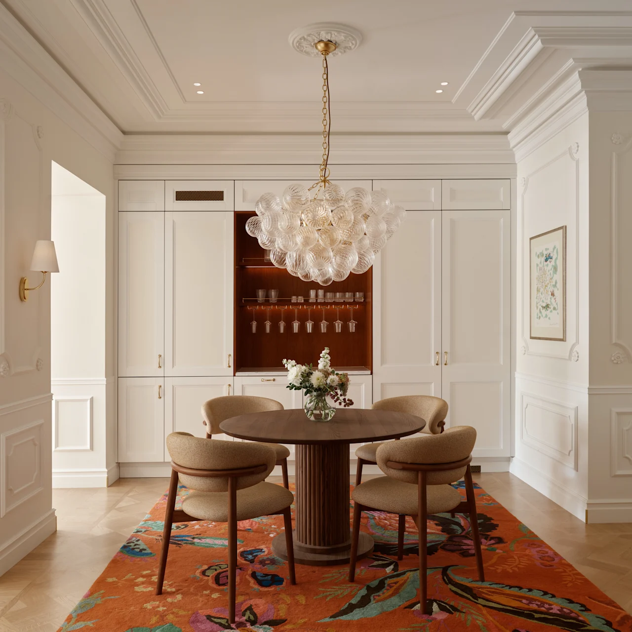 A round wooden table with chairs sits beneath a glass chandelier in an elegant dining room, featuring white paneled walls, built-in cabinets, floral artwork, and a colorful patterned rug.