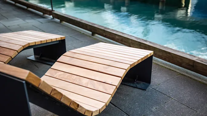 Two wooden curved benches resting on a stone pavement beside a turquoise-colored body of water, surrounded by a wooden railing.